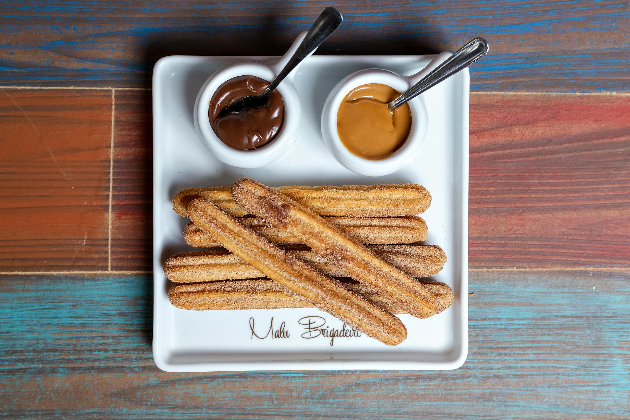 Plate of churros with chocolate and caramel dips on a colorful wooden table.